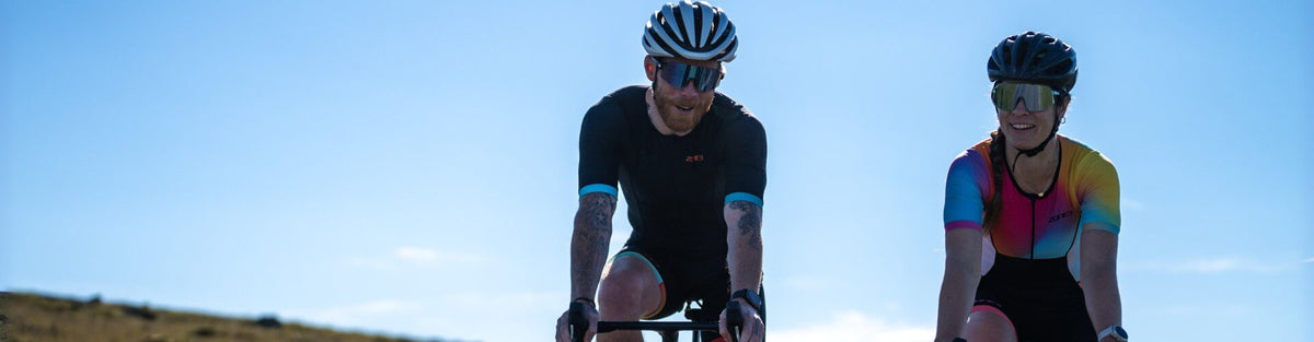 Twee triatleten in ZONE3-trisuits fietsen naast elkaar tegen een blauwe lucht.