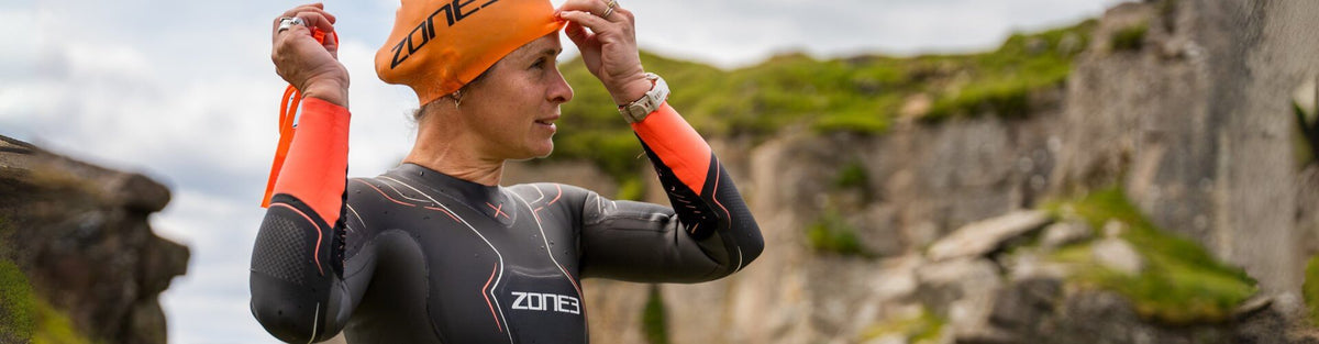 Een zwemmer in een zwarte ZONE3-wetsuit en oranje badmuts past zijn zwembril aan, met groene kliffen op de achtergrond.