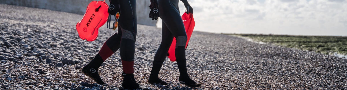 Twee zwemmers in wetsuits lopen over een kiezelstrand met rode ZONE3-sleepboeien.