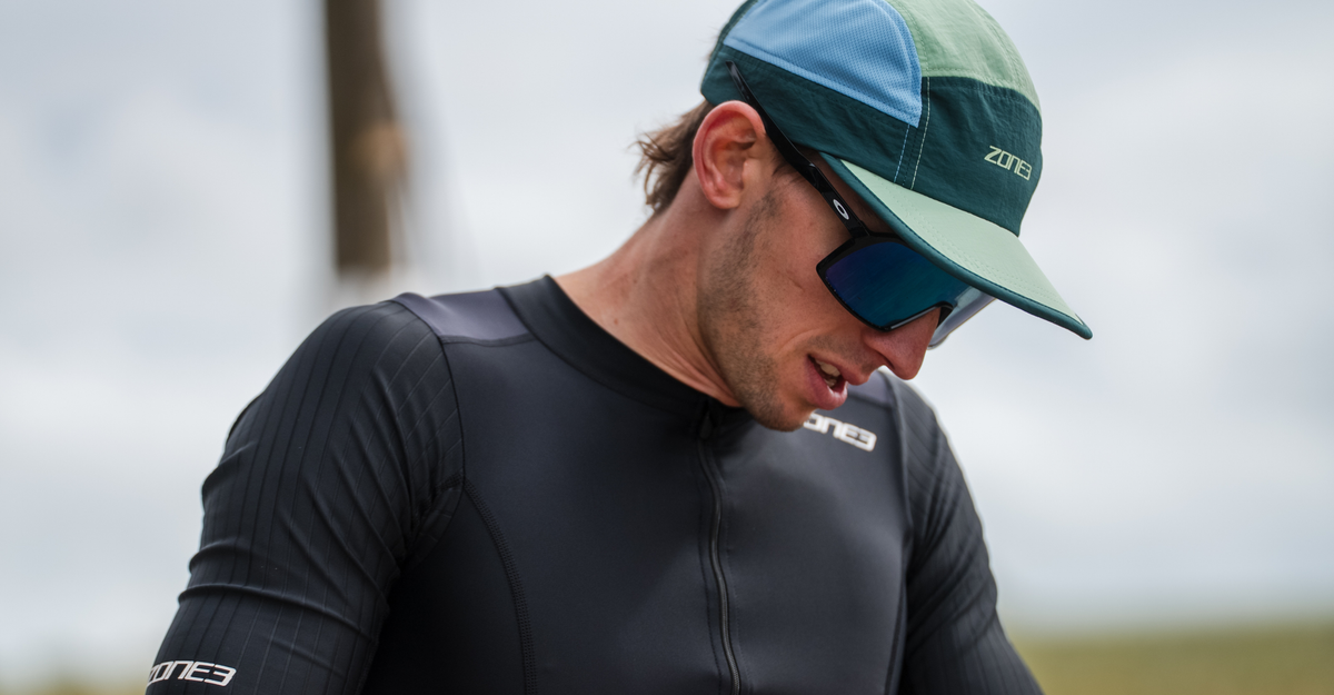 Person wearing black zone3 trisuit with a teal 5 panel running cap and sunglasses, outdoors.