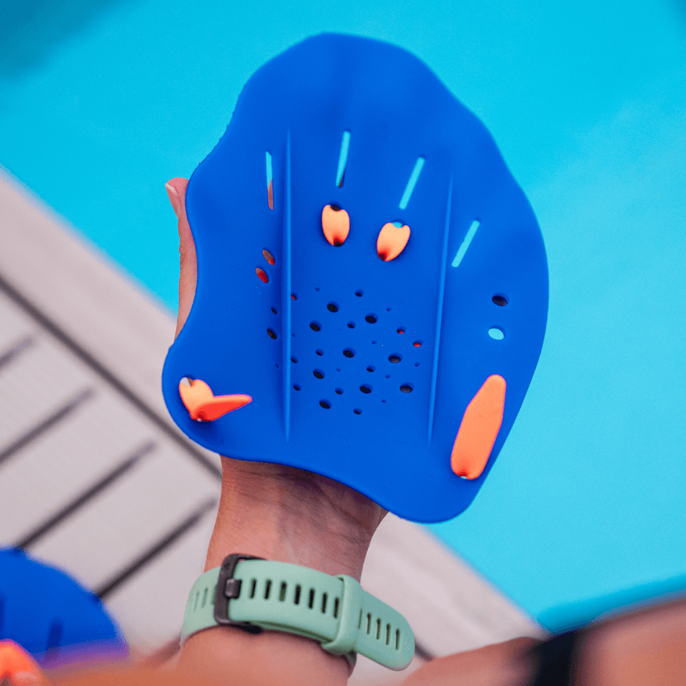 Close-up of blue Ergo Swim Training Hand Paddle with perforations and orange fasteners held by a hand wearing a mint green wristwatch.