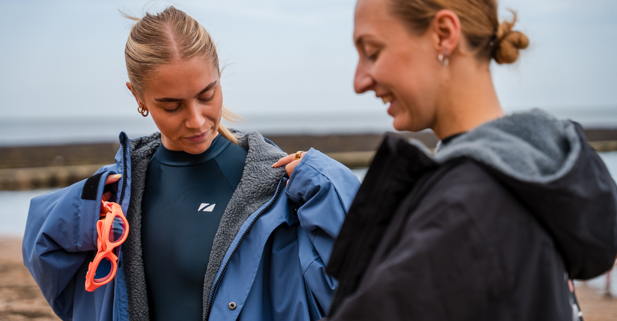 Two women standing outdoors, one wearing a blue jacket with the TI+ thermal swimsuit, against a blurred natural background.