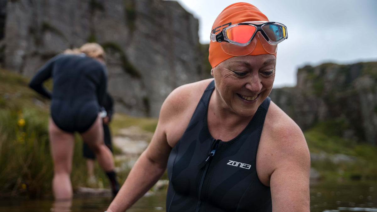 Woman wearing the Zone3 Vision Max Mirror Swim Mask  swim gear and orange swim cap, standing in a natural water body with rocky landscape.