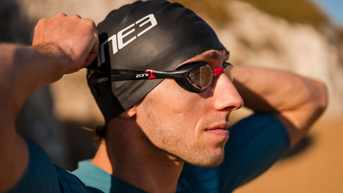 Male Athlete wearing a Zone3 swim cap and Volare Racing goggles with a blurred natural background.