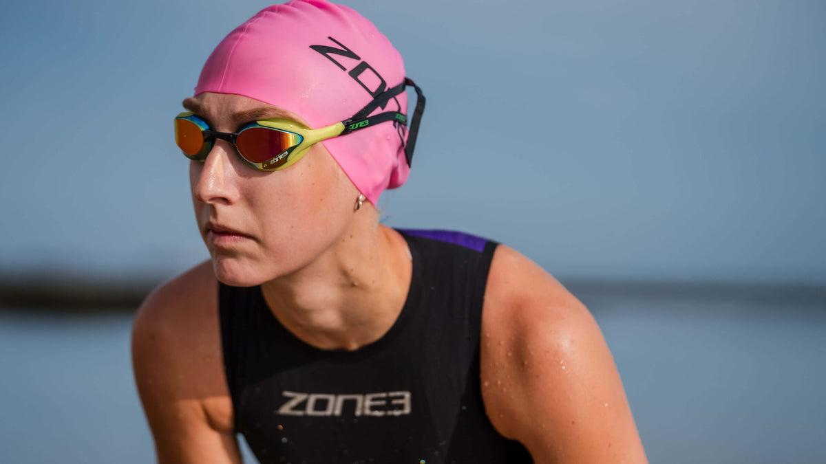 Female Athlete wearing a Zone3 swim cap and Volare Racing goggles with a blurred natural background.