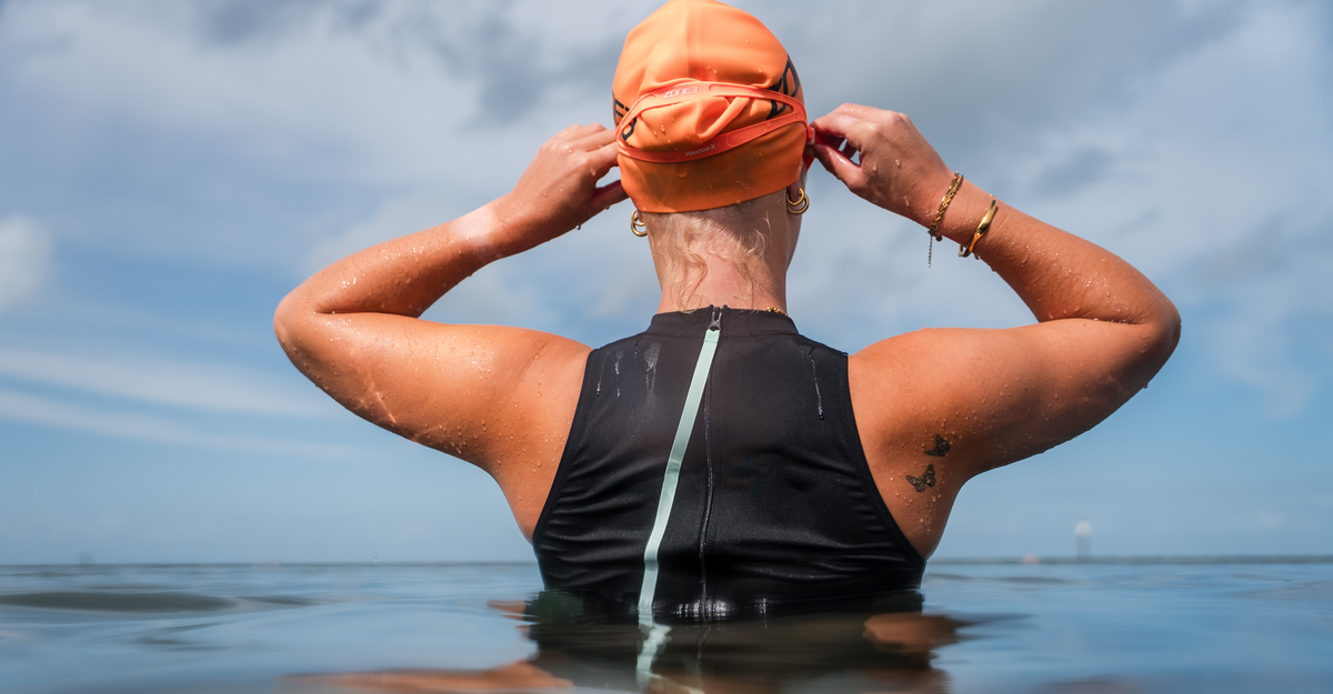 Person wearing an orange swim cap and black high neck swimsuit in water with a blue sky background
