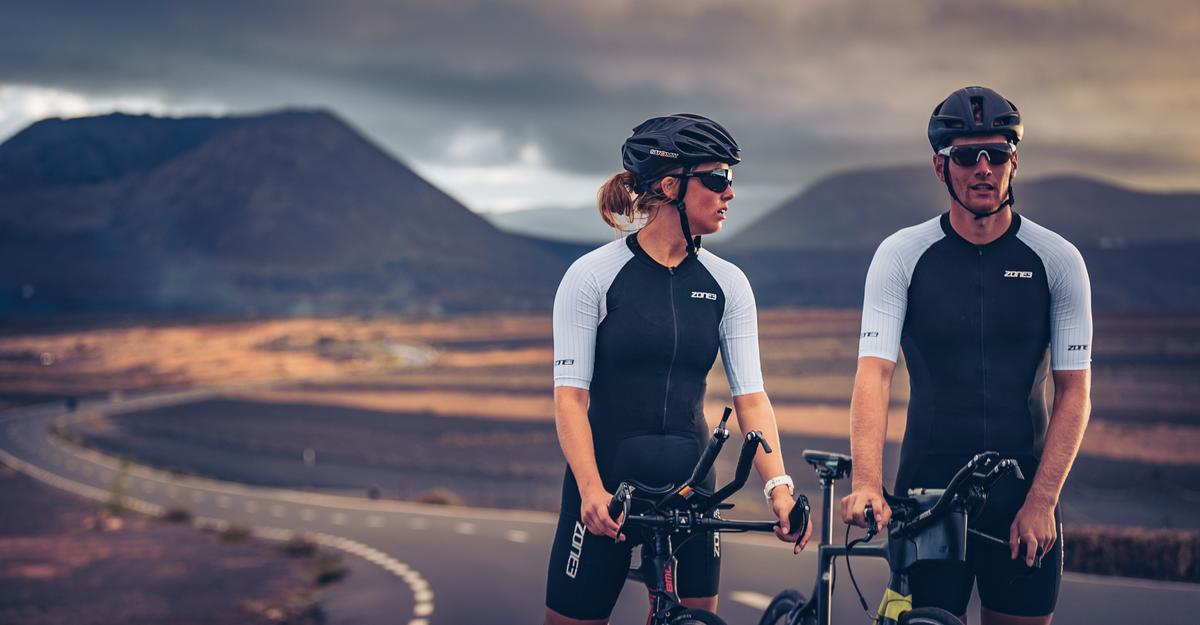 Two cyclists in the lava trisuit  standing on a road with mountains in the background
