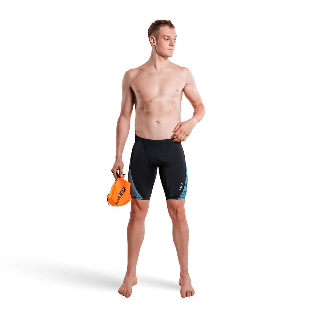 Man wearing black and teal Zone3 Jammers holding an orange swim cap on a white background