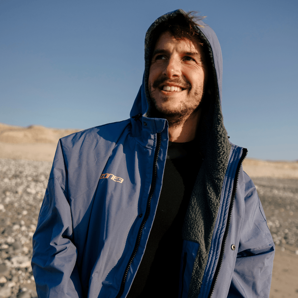 Smiling person wearing the ZONE3 robe partially unzipped, standing on a rocky beach in warm daylight.