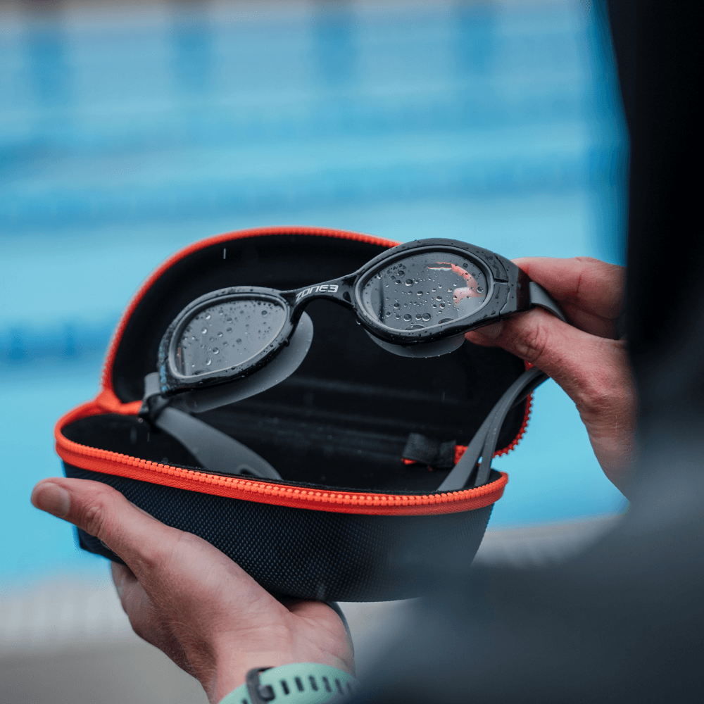 Open navy goggle case showing Zone3 swim goggles with water droplets on the lenses.